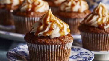 A tray of sticky toffee cupcakes topped with glossy toffee sauce and a swirl of whipped cream, garnished with chopped dates.