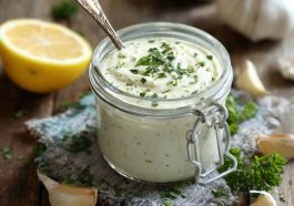 A small bowl of creamy garlic aioli sauce garnished with fresh herbs, surrounded by fries, grilled vegetables, and lemon wedges.