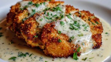 Golden parmesan-crusted chicken served with creamy garlic sauce, garnished with fresh herbs on a rustic plate.