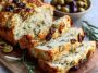 A golden loaf of savory quick bread sliced to reveal chunks of cheese and olives, served with fresh herbs and butter.