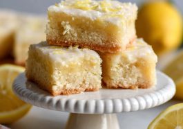 Glazed lemon brownies cut into squares with fresh lemon zest and slices on a serving plate.