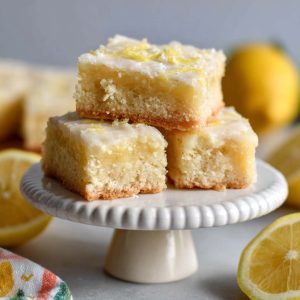 Glazed lemon brownies cut into squares with fresh lemon zest and slices on a serving plate.