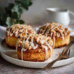 Golden apple cheesecake buns topped with buttery crumble and drizzled with sweet glaze, served on a rustic wooden board.