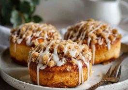 Golden apple cheesecake buns topped with buttery crumble and drizzled with sweet glaze, served on a rustic wooden board.