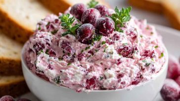 A creamy block of cranberry cream cheese spread topped with ruby-red cranberry sauce and fresh rosemary, served with crackers.