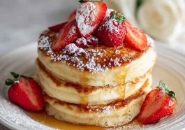 A stack of golden fluffy pancakes topped with melting butter, maple syrup, and fresh berries.
