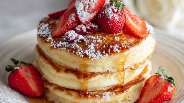 A stack of golden fluffy pancakes topped with melting butter, maple syrup, and fresh berries.