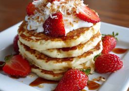 "A stack of fluffy coconut cream pancakes topped with whipped cream, toasted coconut flakes, and fresh fruit."