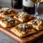 Mini French onion tarts made with golden puff pastry, caramelized onions, and melted Gruyère cheese served on a rustic platter.