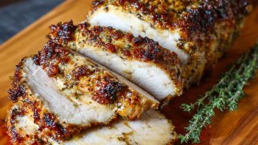 “A golden-brown herb crusted pork loin sliced and served on a rustic wooden board with fresh rosemary and garlic.”