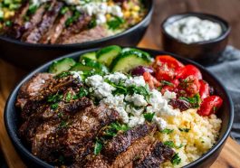 Colorful Mediterranean steak bowl with grilled steak slices, fresh cucumbers, tomatoes, olives, feta cheese, and rice drizzled with tzatziki sauce.