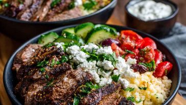 Colorful Mediterranean steak bowl with grilled steak slices, fresh cucumbers, tomatoes, olives, feta cheese, and rice drizzled with tzatziki sauce.