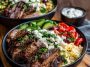 Colorful Mediterranean steak bowl with grilled steak slices, fresh cucumbers, tomatoes, olives, feta cheese, and rice drizzled with tzatziki sauce.