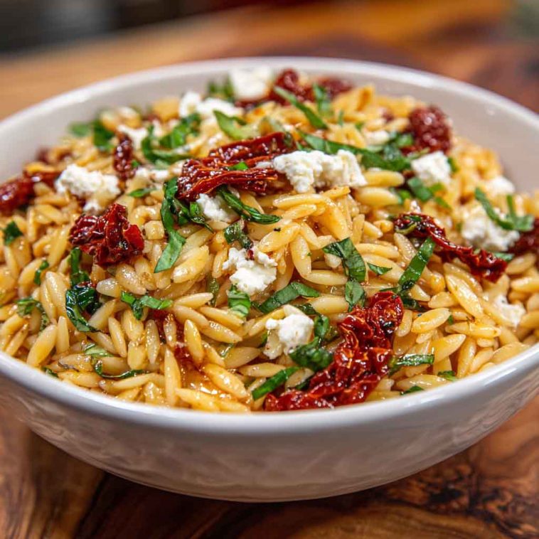 Colorful orzo pasta salad with crumbled feta, sun-dried tomatoes, fresh herbs, and a light Mediterranean dressing served in a bowl.