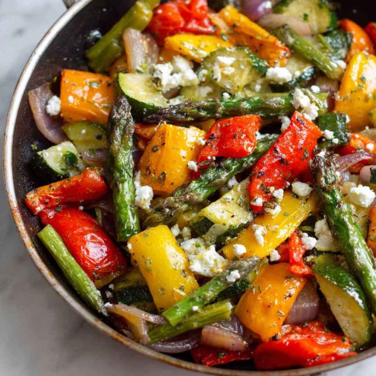 A colorful one-pan dish of roasted Greek vegetables including zucchini, bell peppers, tomatoes, onions, and potatoes, drizzled with olive oil and herbs.