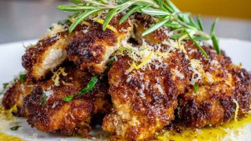 “Golden crispy lemon pecorino chicken served on a plate with fresh herbs and lemon wedges.”