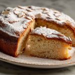 “A golden Italian torta della nonna topped with pine nuts and powdered sugar, sliced to reveal creamy custard filling.”