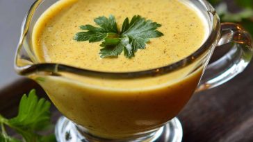 Jar of tangy mustard sauce with a spoon and fresh herbs beside it.