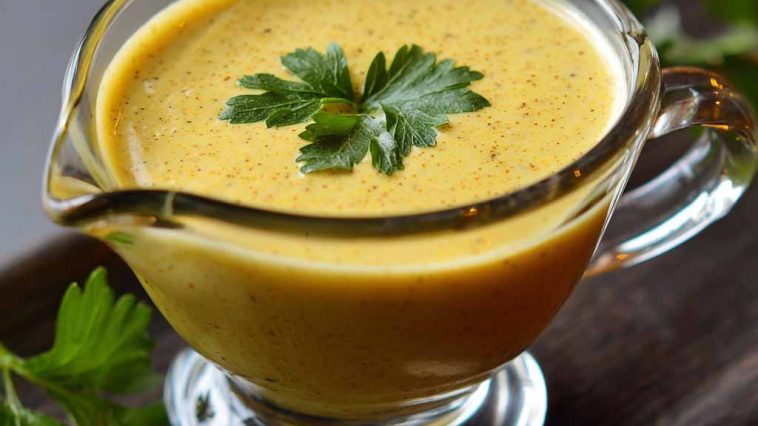 Jar of tangy mustard sauce with a spoon and fresh herbs beside it.