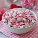 Valentine’s Day muddy buddies coated in pink and red sprinkles, powdered sugar, and chocolate, served in a festive bowl with heart-shaped decorations.