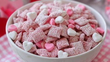 Valentine’s Day muddy buddies coated in pink and red sprinkles, powdered sugar, and chocolate, served in a festive bowl with heart-shaped decorations.