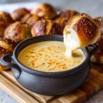 Creamy beer cheese dip served in a bowl, golden and smooth, topped with chives, surrounded by pretzels, chips, and fresh vegetables for dipping.