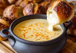 Creamy beer cheese dip served in a bowl, golden and smooth, topped with chives, surrounded by pretzels, chips, and fresh vegetables for dipping.