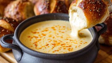 Creamy beer cheese dip served in a bowl, golden and smooth, topped with chives, surrounded by pretzels, chips, and fresh vegetables for dipping.
