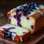 Blueberry cream cheese loaf with golden crust, swirled cream cheese filling, and fresh blueberries, sliced and served on a wooden board.