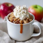 Protein apple crisp in a mug topped with golden oat crumble, warm cinnamon apples, and a dollop of Greek yogurt, served as a quick single-serve dessert.