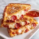 Pizza grilled cheese sandwich with golden toasted bread, melted mozzarella, pepperoni slices, and marinara sauce, served hot with a dipping cup of tomato sauce.