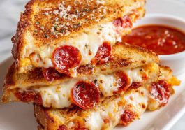 Pizza grilled cheese sandwich with golden toasted bread, melted mozzarella, pepperoni slices, and marinara sauce, served hot with a dipping cup of tomato sauce.