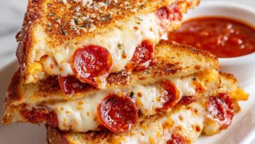 Pizza grilled cheese sandwich with golden toasted bread, melted mozzarella, pepperoni slices, and marinara sauce, served hot with a dipping cup of tomato sauce.