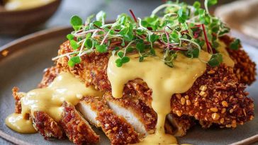 Crispy pretzel chicken served with creamy mustard-cheddar sauce, golden brown breaded chicken cutlets topped with rich cheese sauce, garnished with parsley and served on a rustic plate.