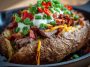 Loaded baked potato topped with juicy grilled steak slices, melted cheese, sour cream, crispy bacon bits, and fresh chives, served on a rustic plate.