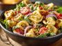 Colorful tortellini salad with cherry tomatoes, olives, fresh herbs, and shredded cheese, served in a large salad bowl.