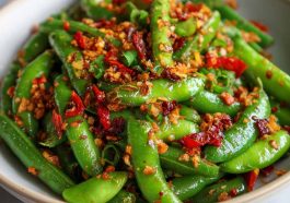 Crisp snap peas tossed in spicy chili crisp sauce with sesame seeds and fresh herbs, served in a white salad bowl.