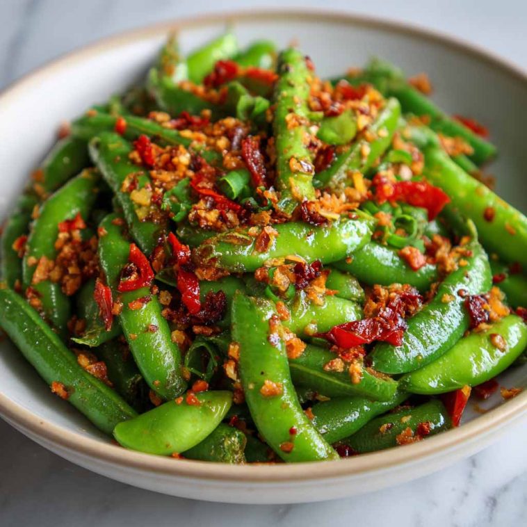 Crisp snap peas tossed in spicy chili crisp sauce with sesame seeds and fresh herbs, served in a white salad bowl.