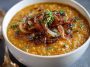A warm bowl of caramelized onion red lentil soup with a rich golden color, topped with herbs and served with crusty bread.