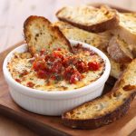 Bubbly three‑cheese tomato bruschetta dip baked in a skillet, topped with fresh basil and served with toasted bread slices.