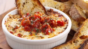 Bubbly three‑cheese tomato bruschetta dip baked in a skillet, topped with fresh basil and served with toasted bread slices.