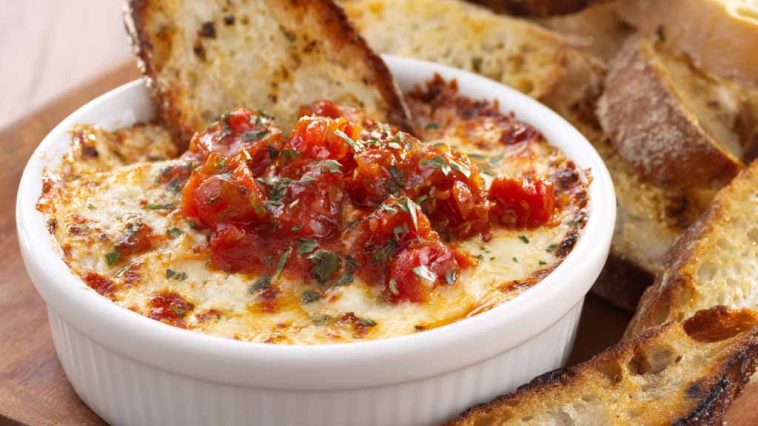 Bubbly three‑cheese tomato bruschetta dip baked in a skillet, topped with fresh basil and served with toasted bread slices.