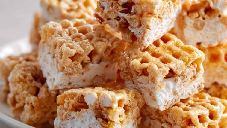 Chewy marshmallow cereal treats made with honeycomb cereal, cut into squares and stacked on a plate with a gooey texture.