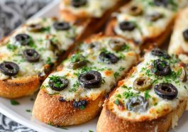 Toasted French bread topped with melted cheese, sliced olives, and herbs, served as a savory appetizer.