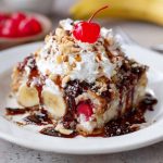 Banana split dump cake topped with melted chocolate, cherries, and whipped cream served in a baking dish.