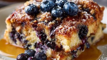 A golden-baked blueberry French toast casserole topped with fresh blueberries, powdered sugar, and drizzled with syrup, served in a baking dish.