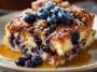 A golden-baked blueberry French toast casserole topped with fresh blueberries, powdered sugar, and drizzled with syrup, served in a baking dish.
