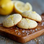 A plate of golden lemon sugar cookies sprinkled with sugar crystals, garnished with lemon zest and served on a rustic white dish.