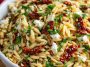 A colorful bowl of orzo pasta salad with crumbled feta, sun-dried tomatoes, fresh herbs, and a light vinaigrette, served in a rustic dish.