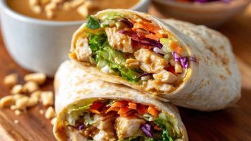 Fresh Thai peanut chicken wraps filled with grilled chicken, crunchy vegetables, and drizzled with creamy peanut sauce, served on a wooden board.